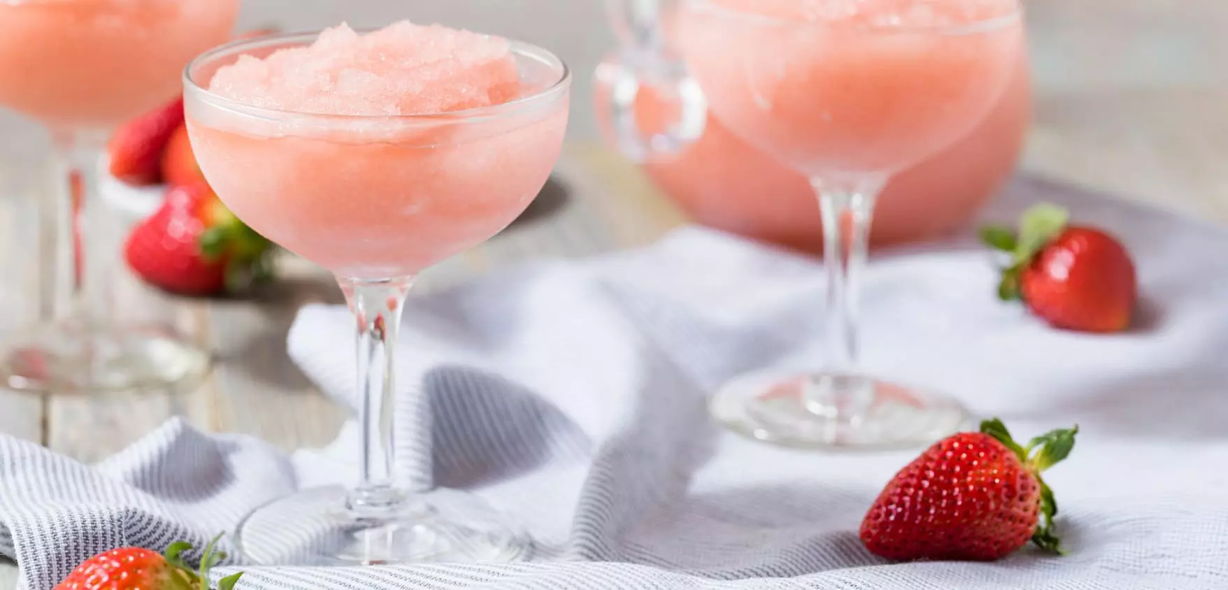Pink frozen drink in coupe glasses with fresh strawberries on a cloth napkin.