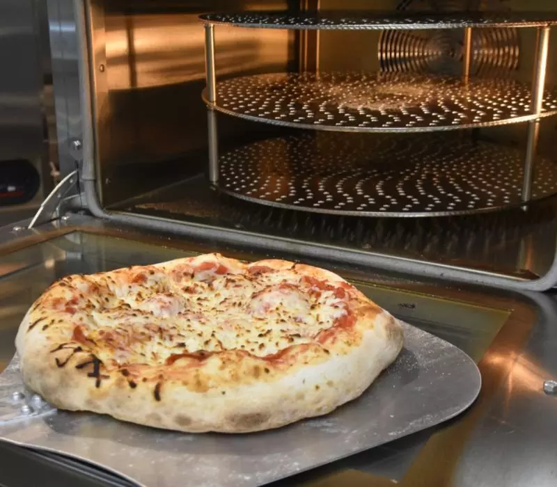 Pizza on a metal peel in front of an open industrial pizza oven.