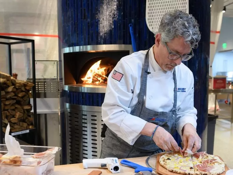 Chef in white uniform preparing pizza near a wood-fired oven.