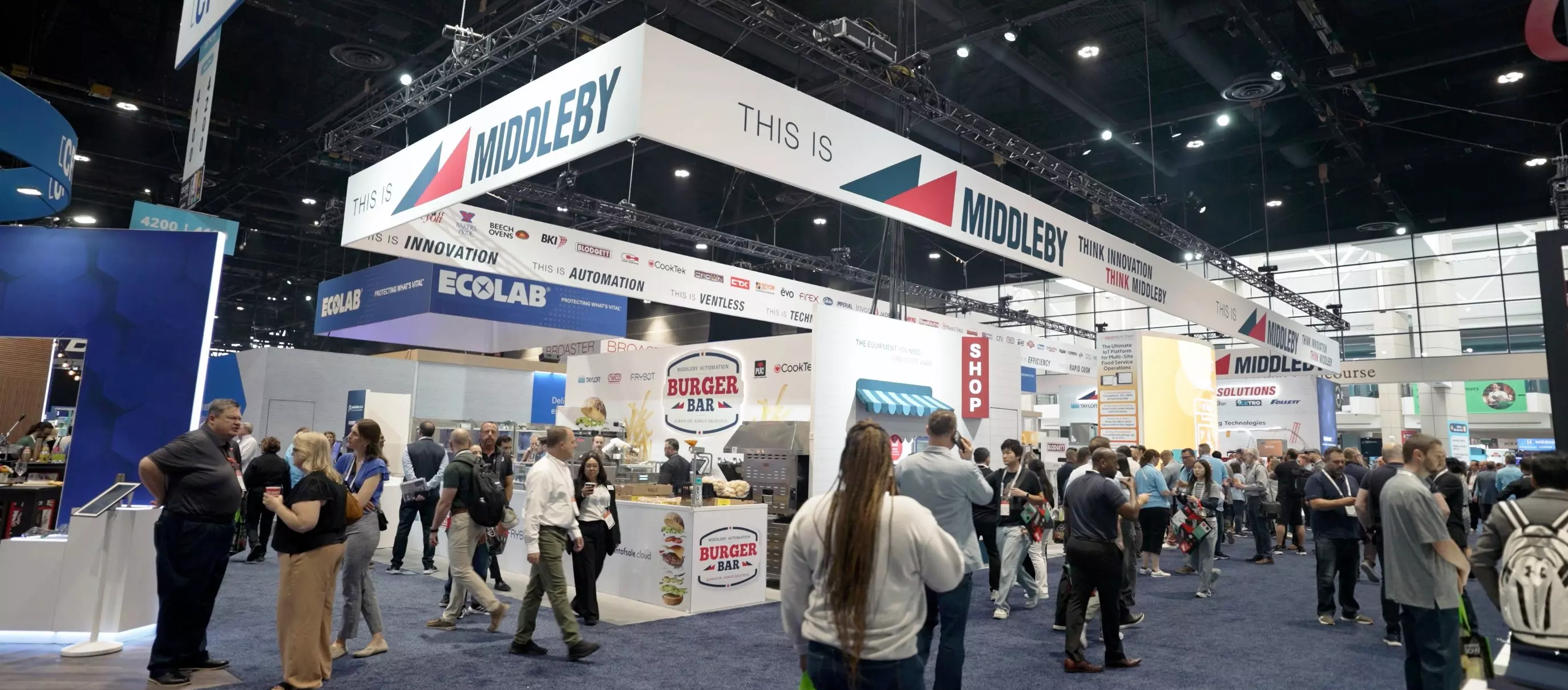 Crowd at Middleby trade show booth with hanging Middleby signs and a Burger Bar demo area.
