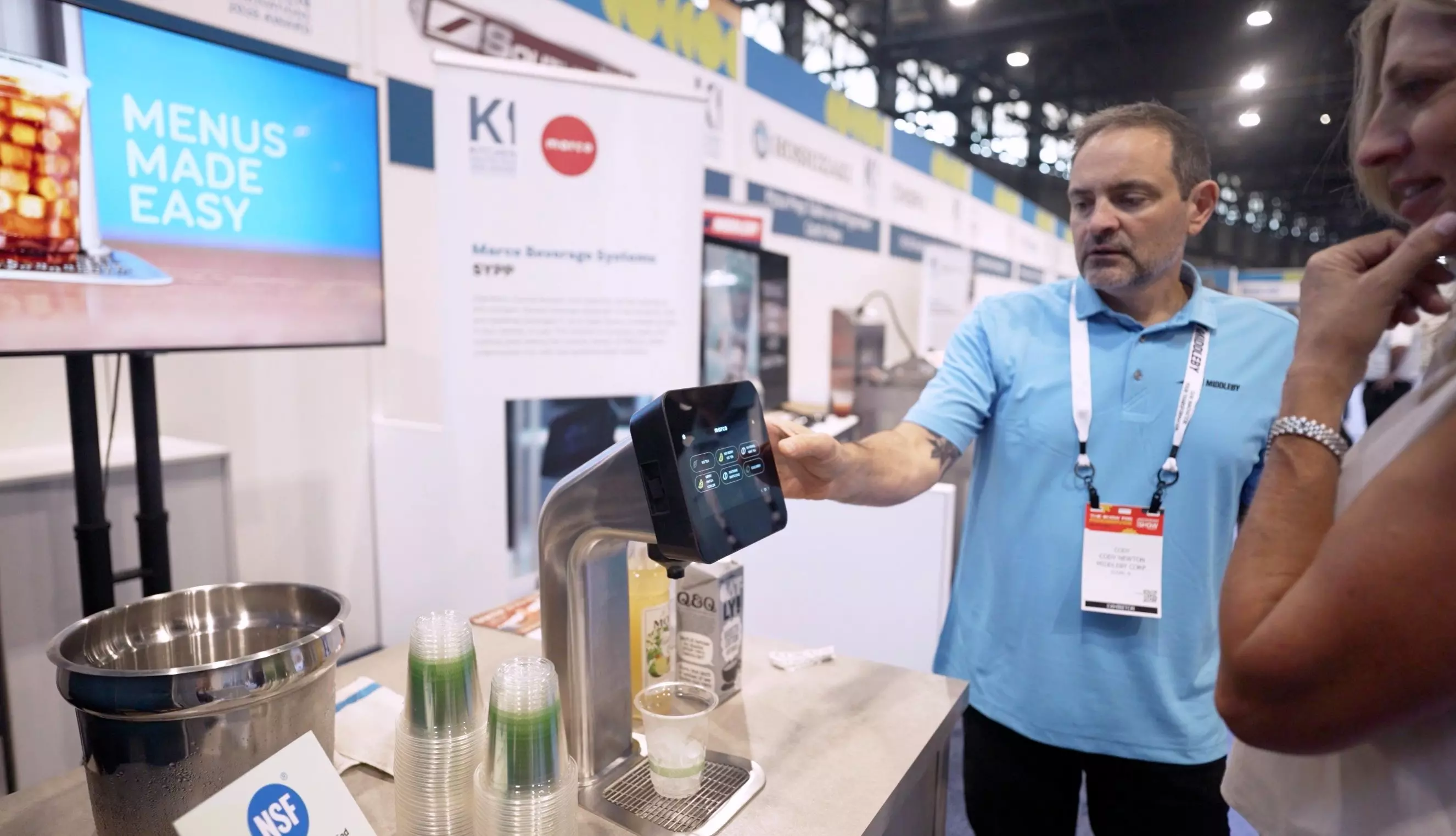 Man demonstrates a touchscreen beverage dispenser at a trade show booth.