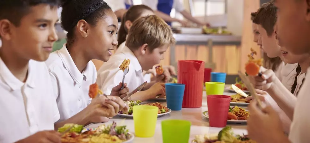 A photo showing a group of students eating together in a cafeteria with a well-balanced meal on their plates as part of their school nutrition program
