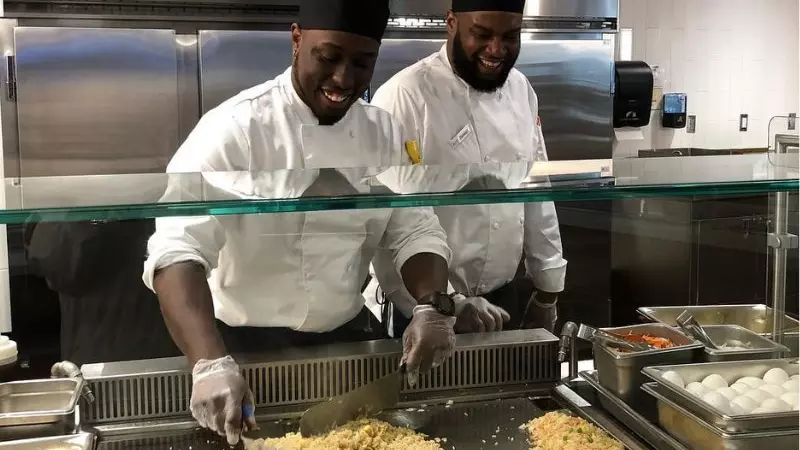 Two chefs in white uniforms cooking fried rice on a griddle, smiling.