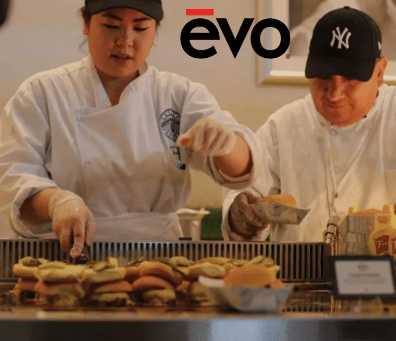 Two chefs assemble burgers on a grill. evo logo visible above.
