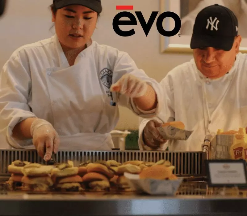 Two chefs assemble burgers on a grill. evo logo visible above.