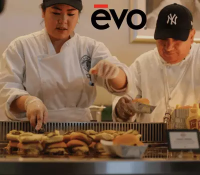 Two chefs assemble burgers on a grill. evo logo visible above.