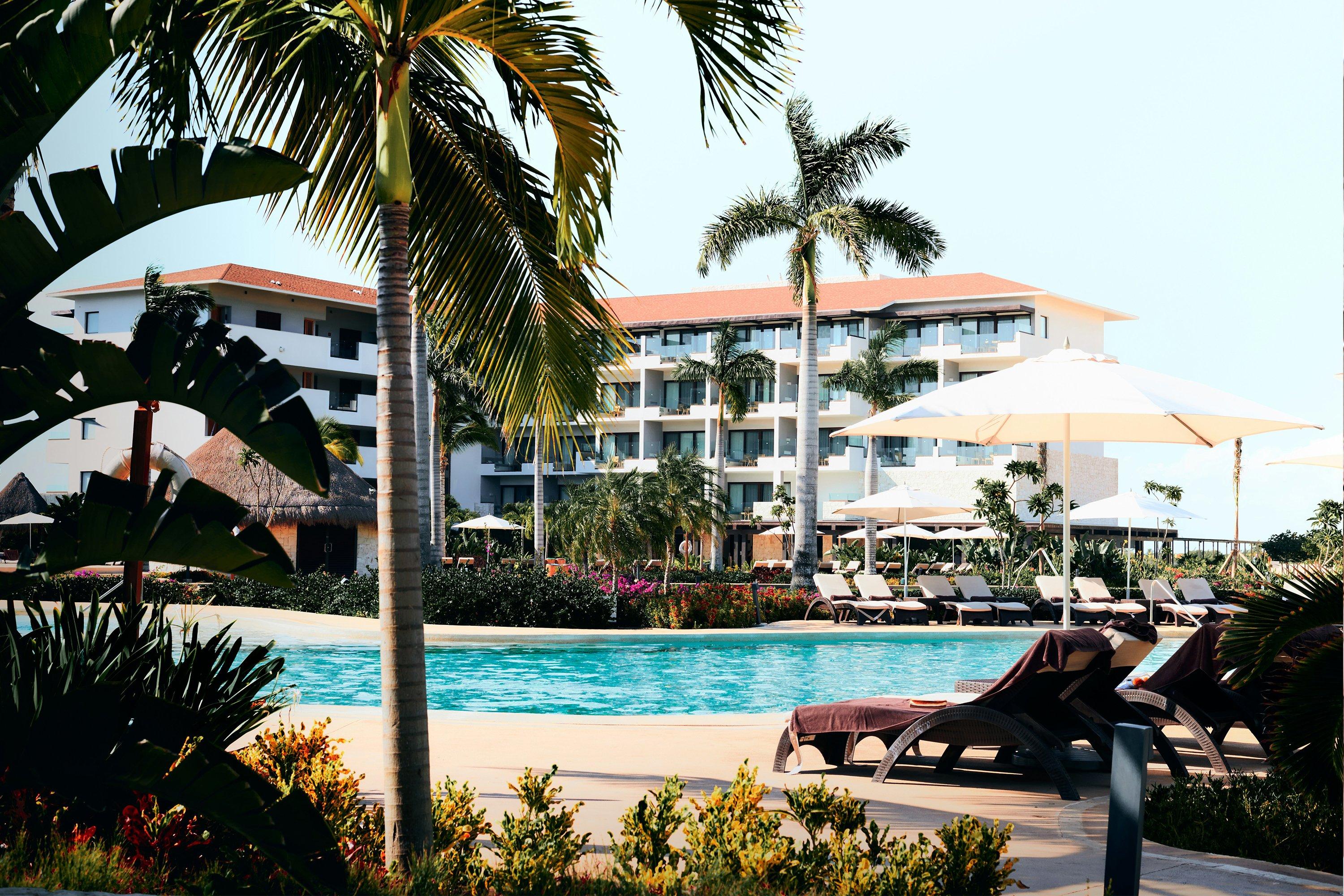 a pool and lounge chairs near a resort hotel with palm trees