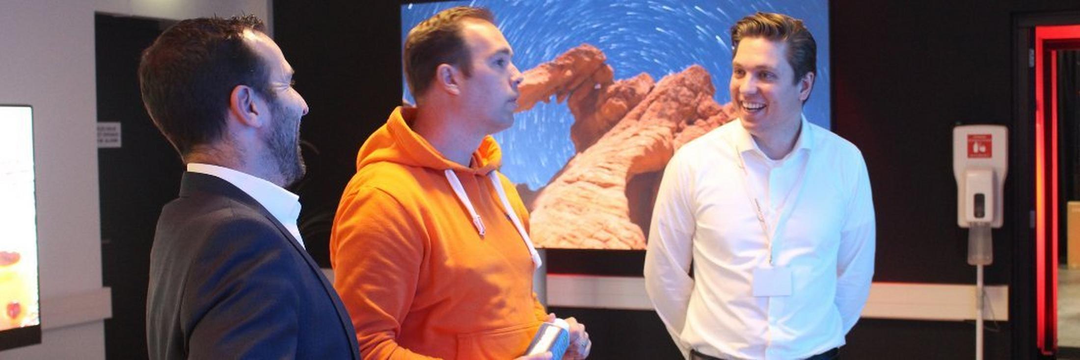 three men standing in front of a large screen with a man in a suit