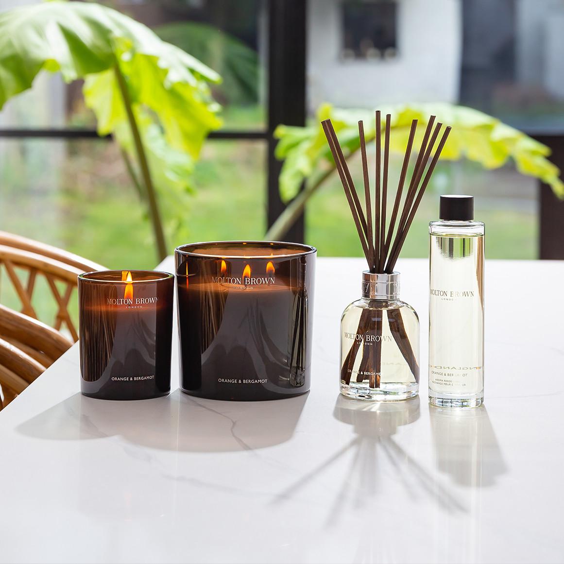 Molton Brown Orange & Bergamot candles and reed diffuser displayed on a white table by a window.