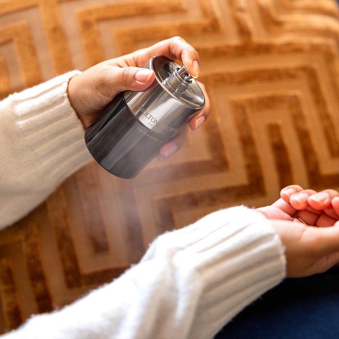 Hand spraying Molton Brown perfume onto wrist, chrome bottle over patterned wooden floor