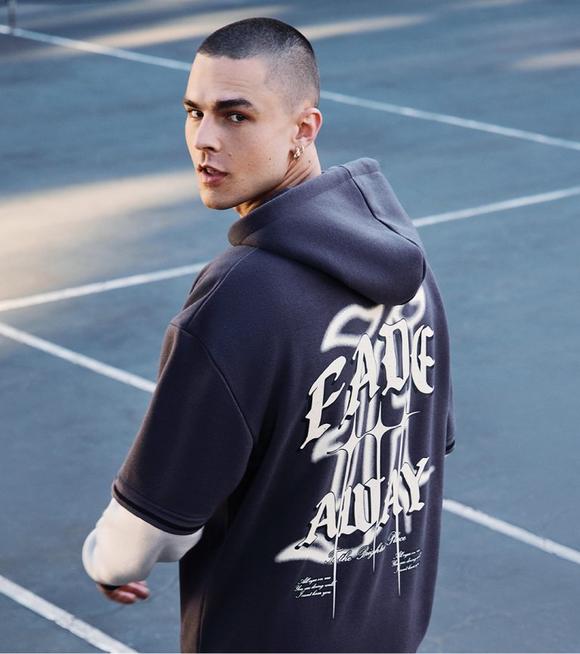 Young man in a hooded sweatshirt with 'fade away' printed on the back, standing in a parking lot, looking over his shoulder.