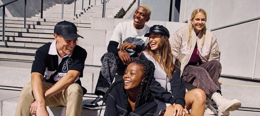 Five young adults laughing and sitting together on concrete steps outdoors, dressed in casual, trendy outfits