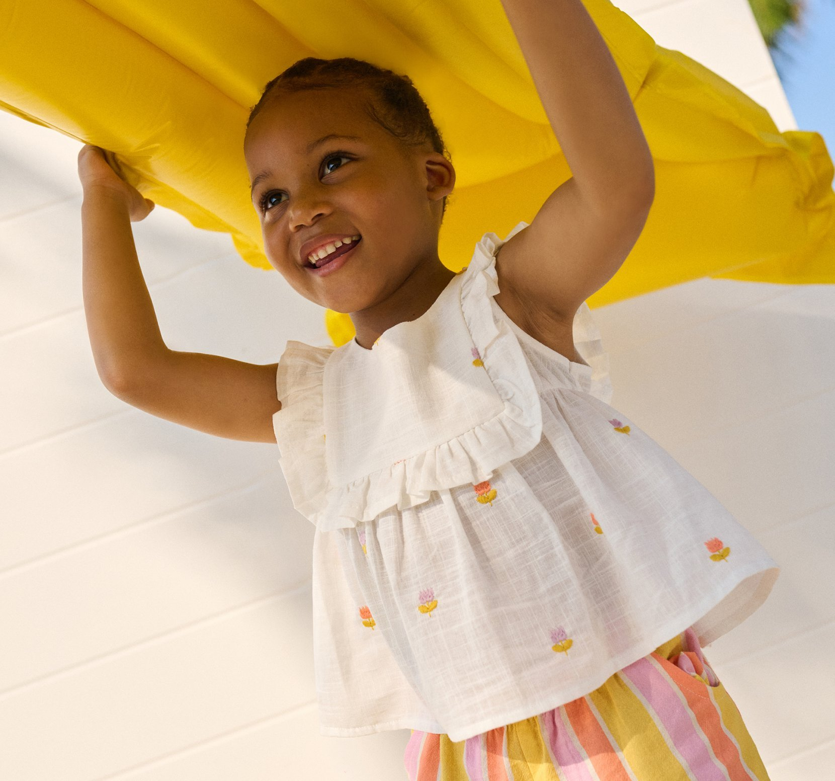 a baby wearing a pattern dress with pink, yellow and blue birds holding a toy