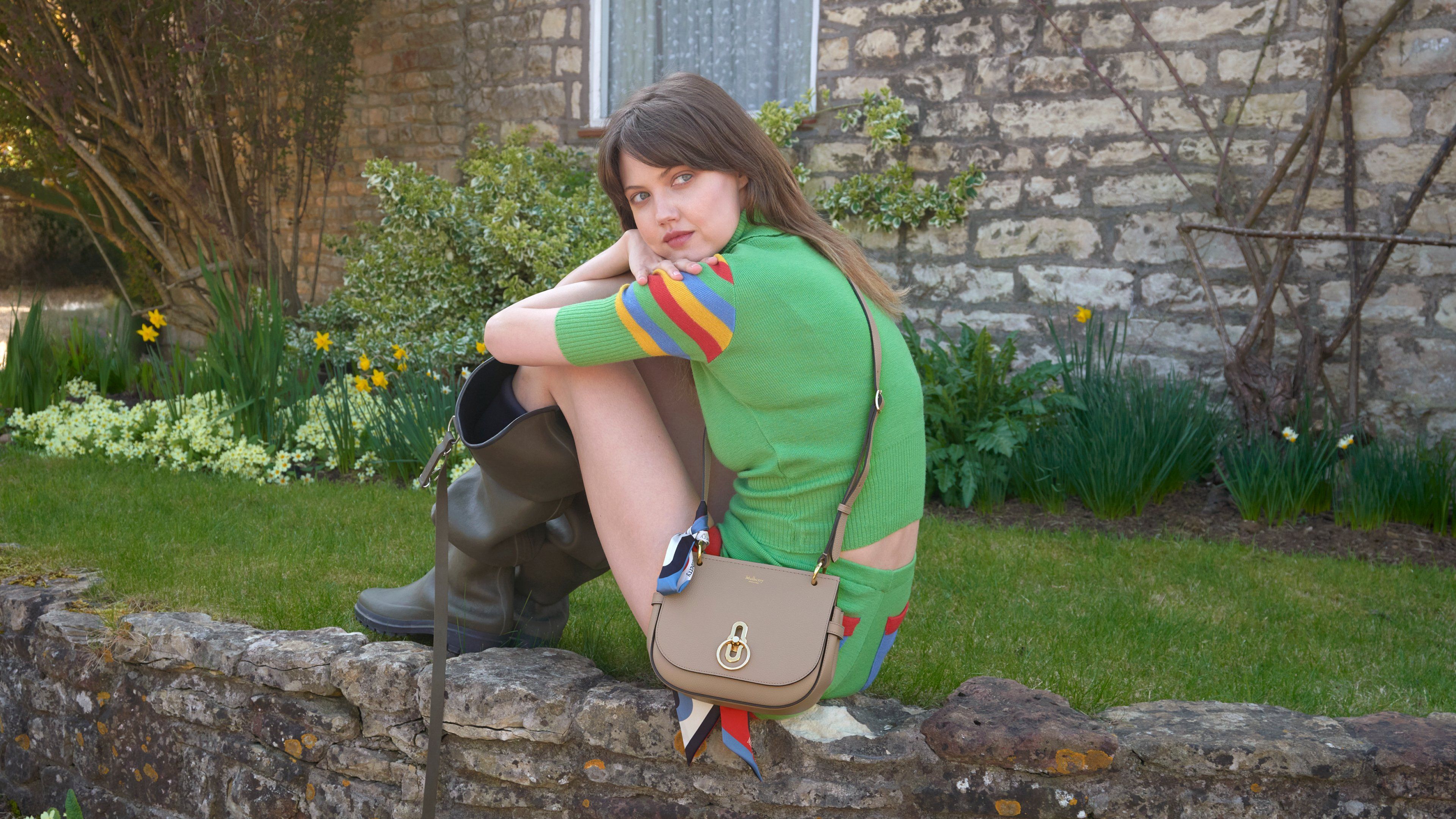 Model sitting on a stone wall wearing the Mulberry Small Amberley handbag in beige leather