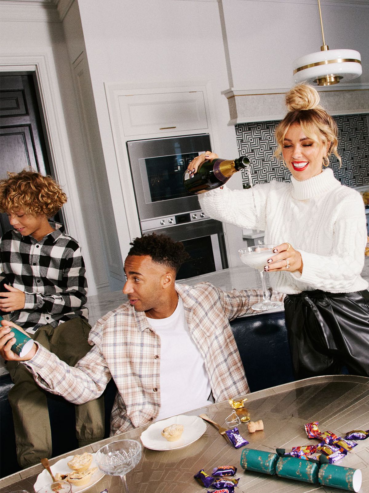Two parents and a child model are seen at a table with a kitchen in the background, the model parent on the right is pouring a glass of prosecco while the parent on the left is holding a cracker with a person out of shot