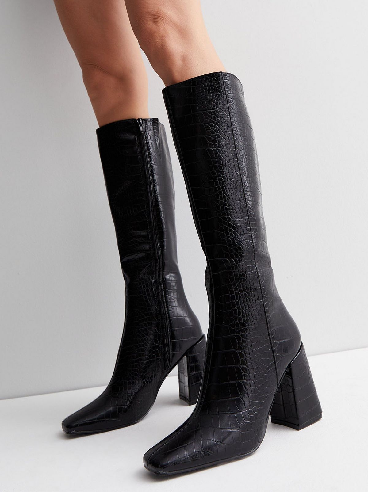A model stands in front of a white background, camera focused on their shoes, they are wearing a pair of black faux croc block heel knee high boots
