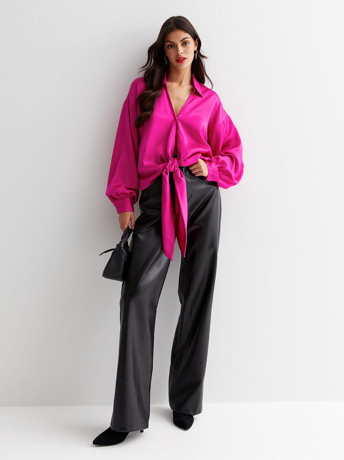 A model stands in front of a white background wearing a bright pink long sleeve collared top and a pair of faux leather trousers