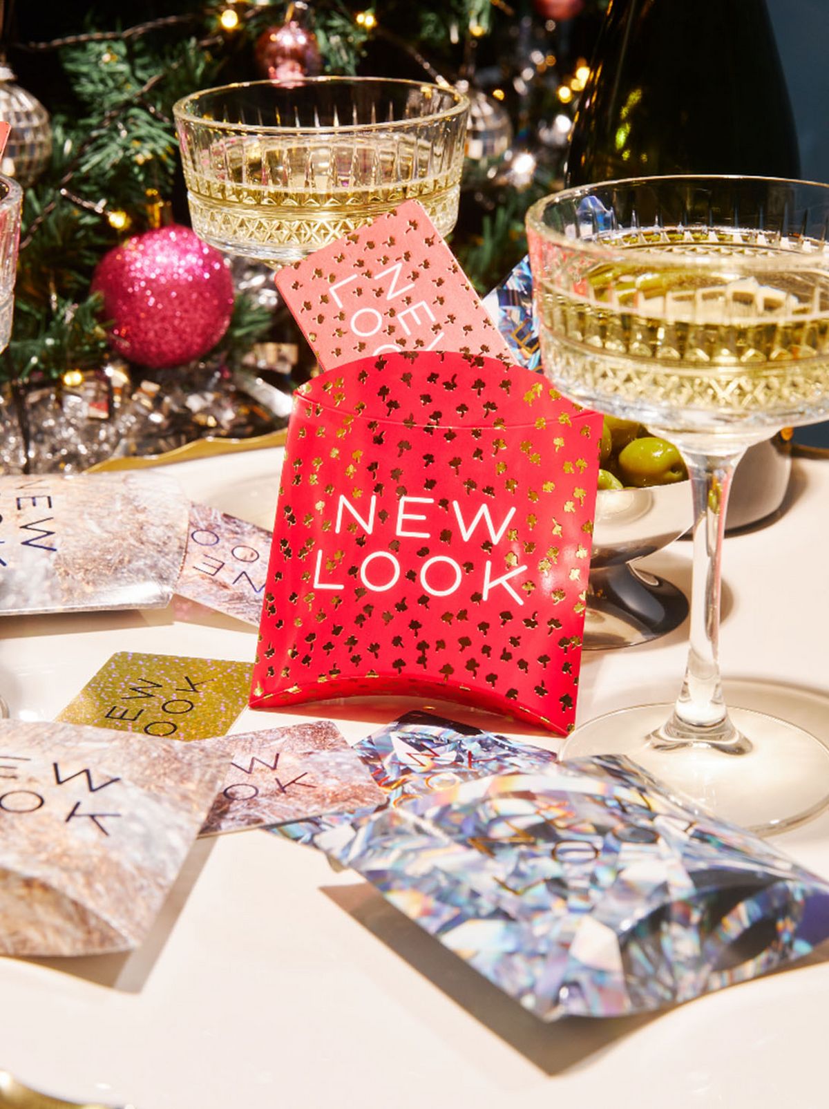 A red gift card with New Look in white on the front is placed in the center of a festive table, with flutes of drink positioned either side and a decorated Christmas tree in the background