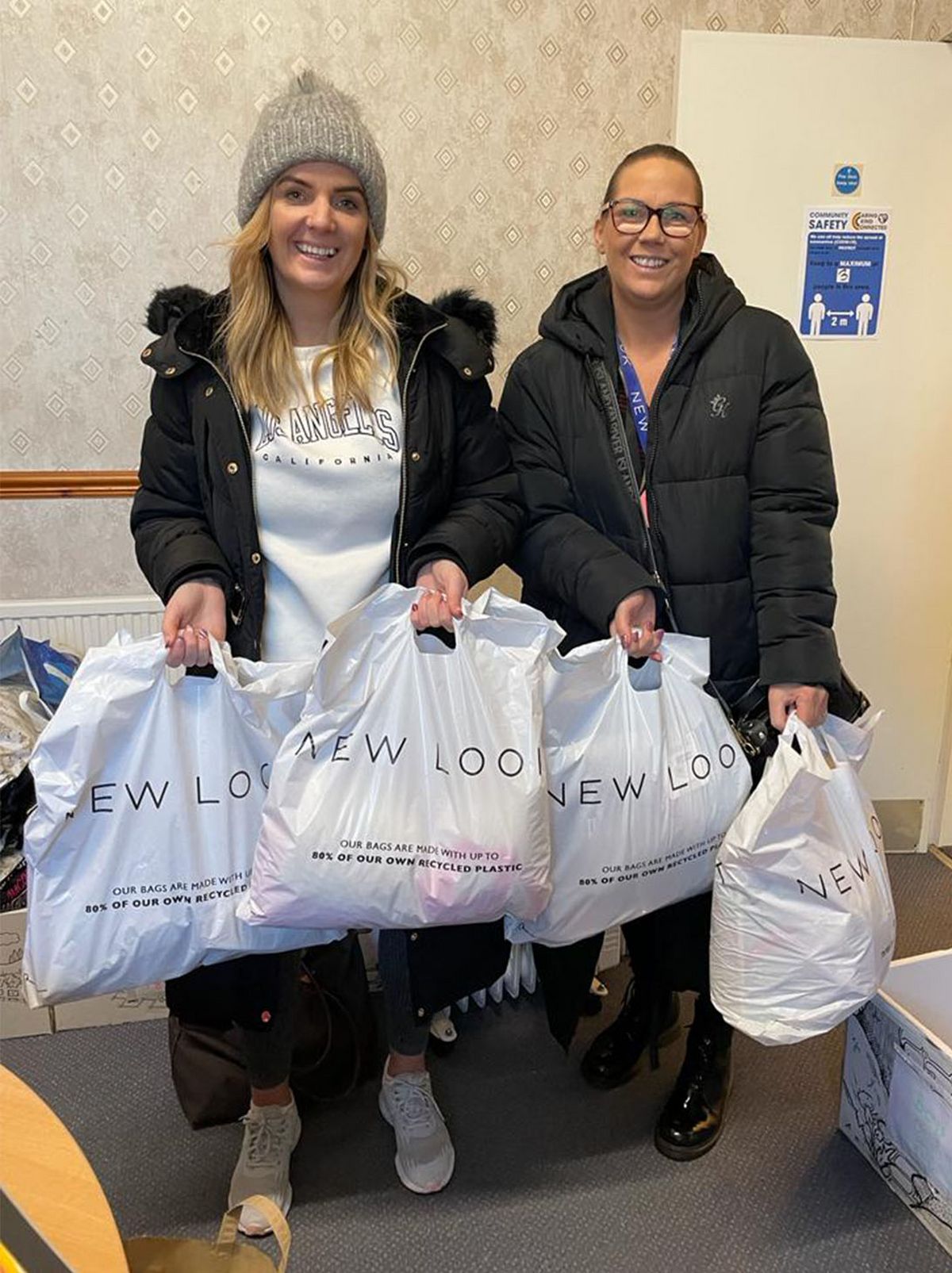 Two people stand in a room smiling at the camera while holding white New Look plastic bags in each hand