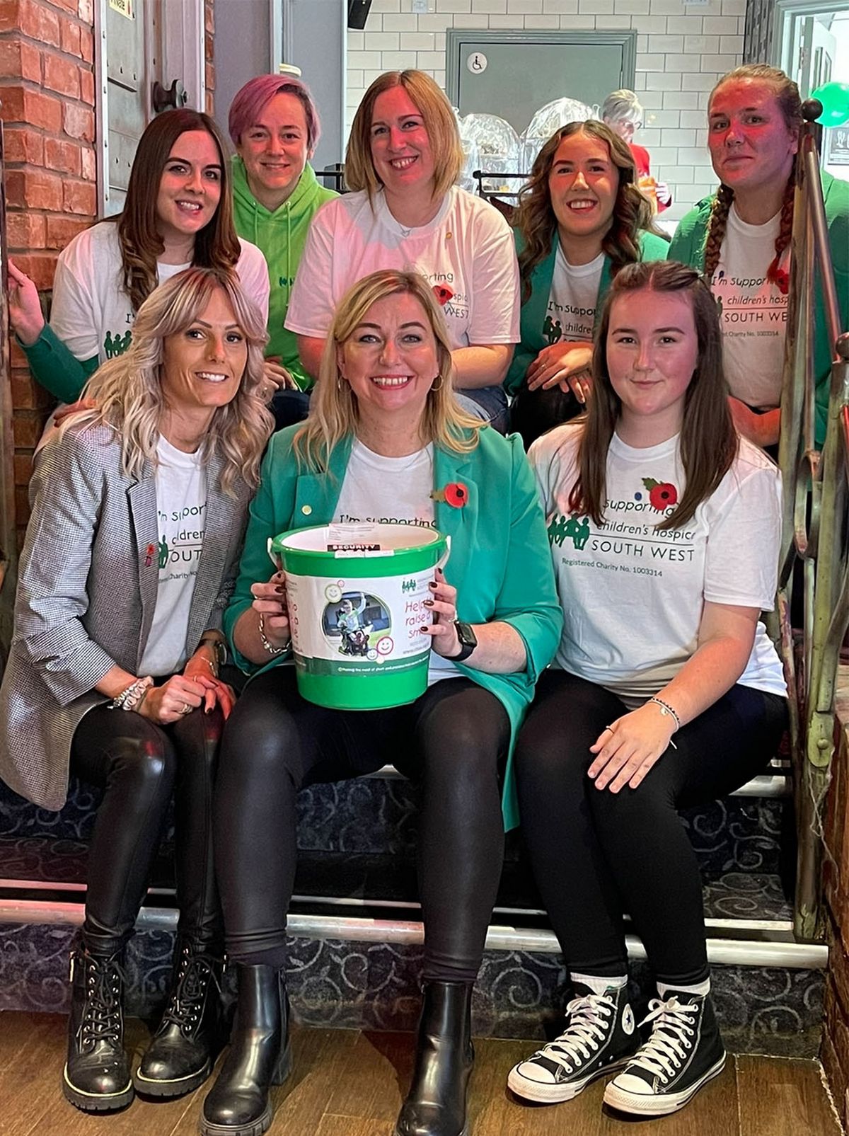 A group of people are sitting on steps smiling at the camera, with the person in the middle holding a large charity bucket on their lap