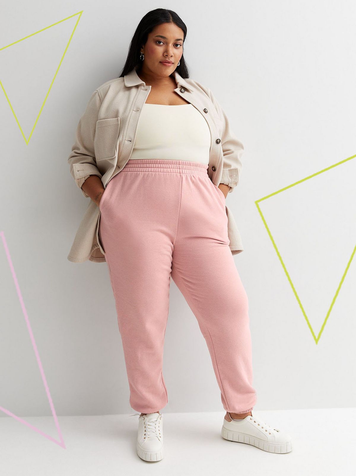 A model leans against a white wall looking directly at the camera with their hands in their pockets, wearing a beige jacket, a white top and a pair of pale pink cuffed joggers