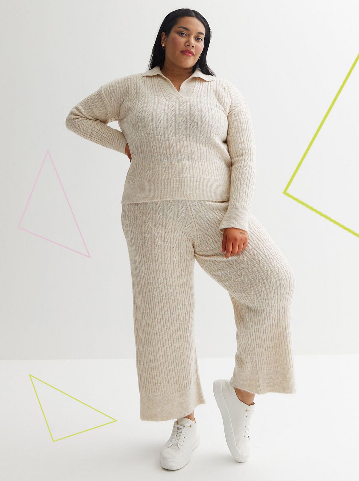 A model stands with a hand on their hip looking directly at the camera on a white wall in front of a white background, they are wearing a pair of cream cable knit wide leg trousers and a matching sweatshirt
