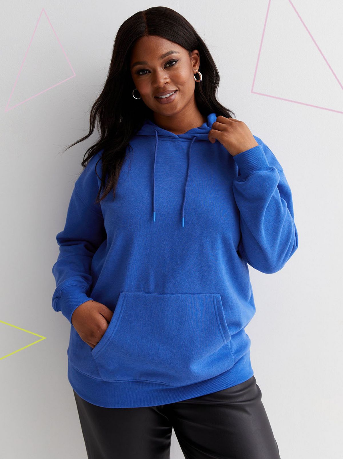 A model stands looking straight at the camera in front of a white background touching their hair with a hand in their jumper pocket, they are wearing a pair of leather look trousers and a bright blue hoodie