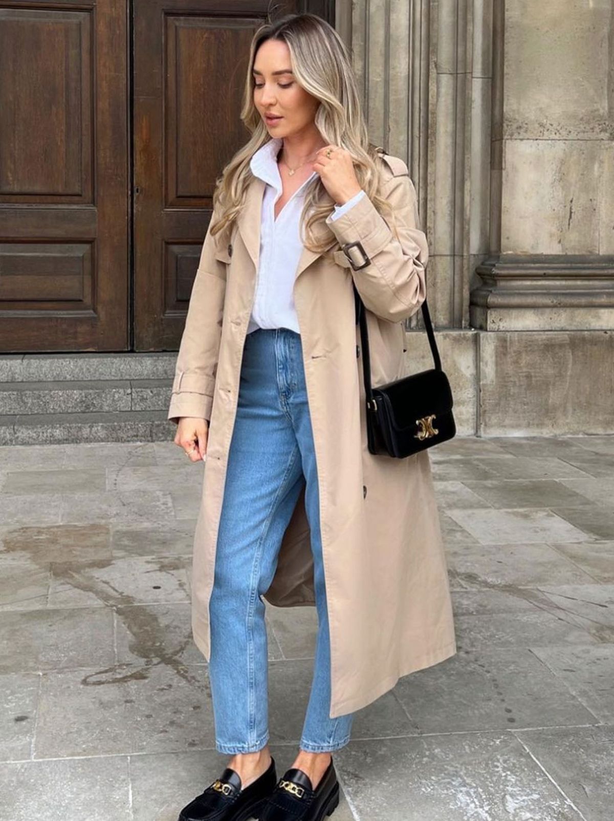 A model stands in front of a large building wearing a long cream jacket, a shirt and a pair of jeans