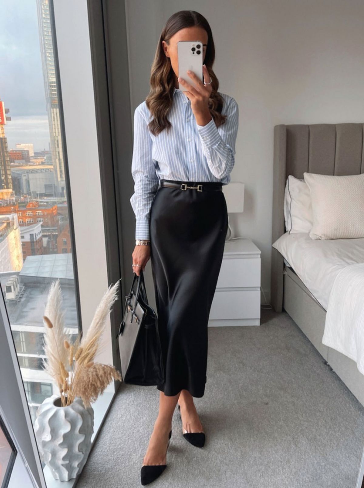 A model stands in a bedroom wearing a blue shirt and a long black skirt