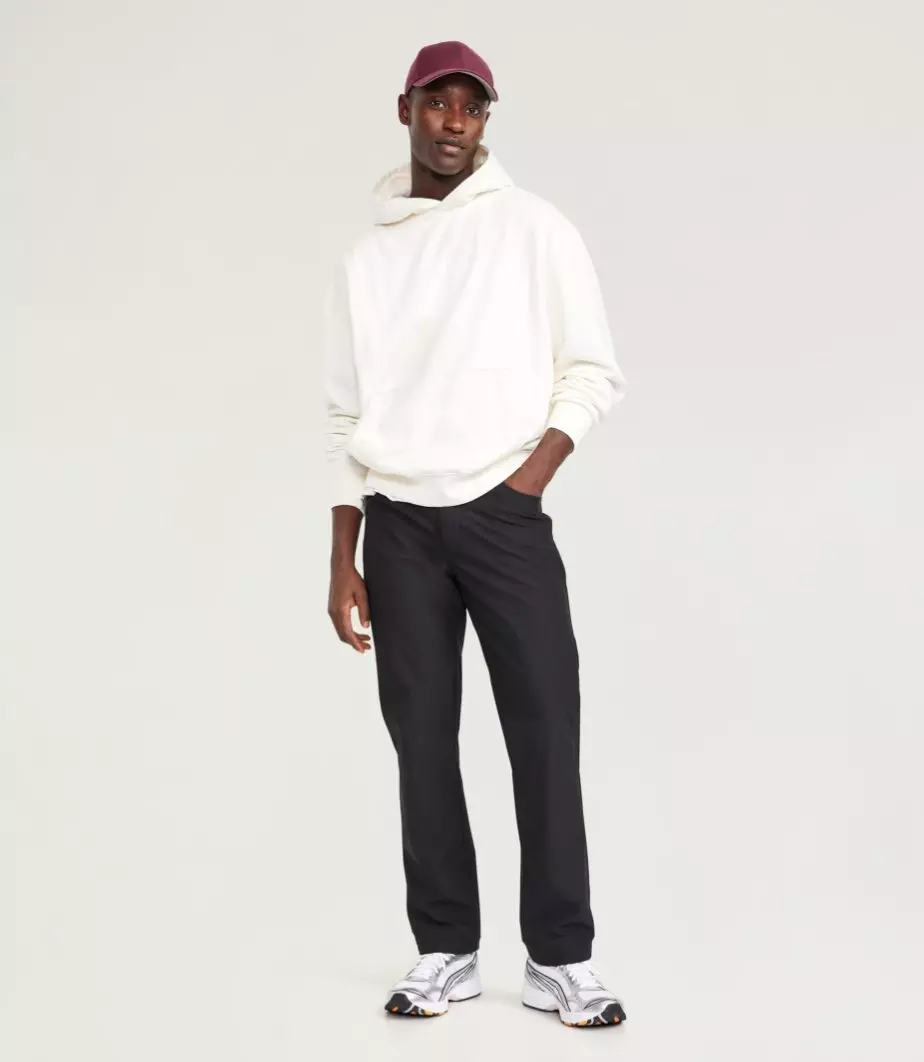 A male model wears dark active pants, a cream sweatshirt, and baseball cap.