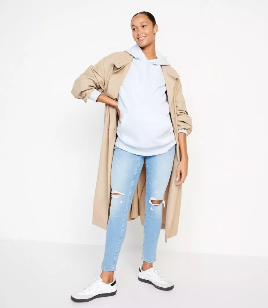 A pregnant model wearing a grey hoodie with a tan trench coat and distressed jeans and sneakers. 