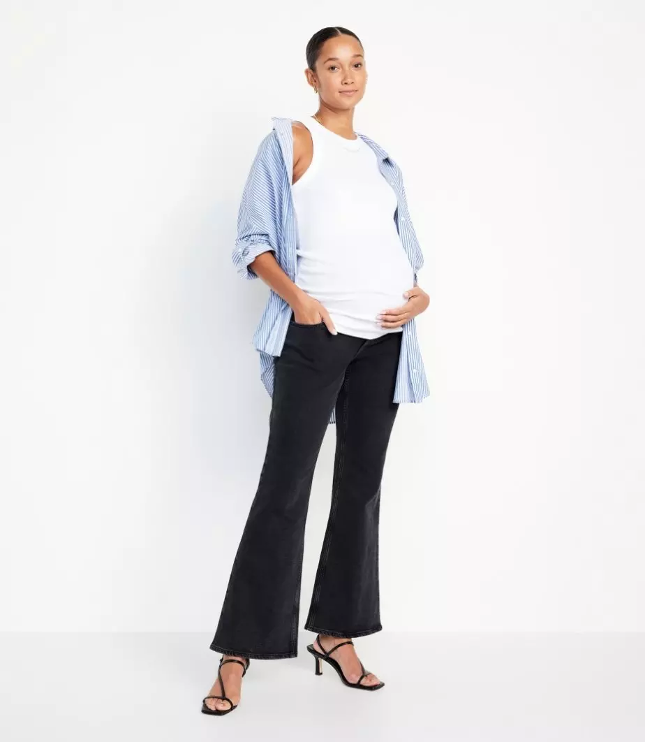Une femme enceinte vêtue d’une camisole de maternité blanche, d’un chemisier bleu et d’un jean à jambe large noir.