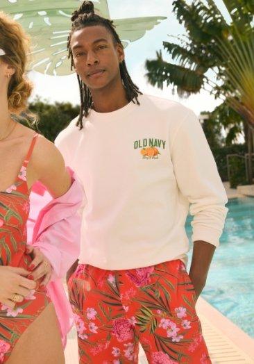 A male model wearing white graphic crew-neck sweatshirts and floral print swim trunks.