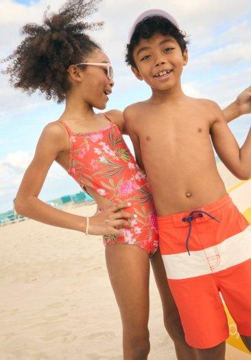 A young girl model wearing a printed one-piece swimsuit and a young boy model wearing orange stripe trunks.