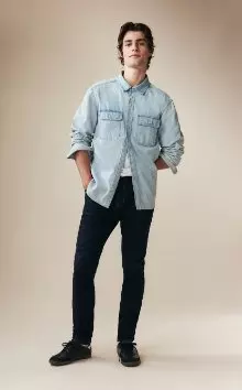 A male model wearing skinny jeans and a light denim button-down shirt.