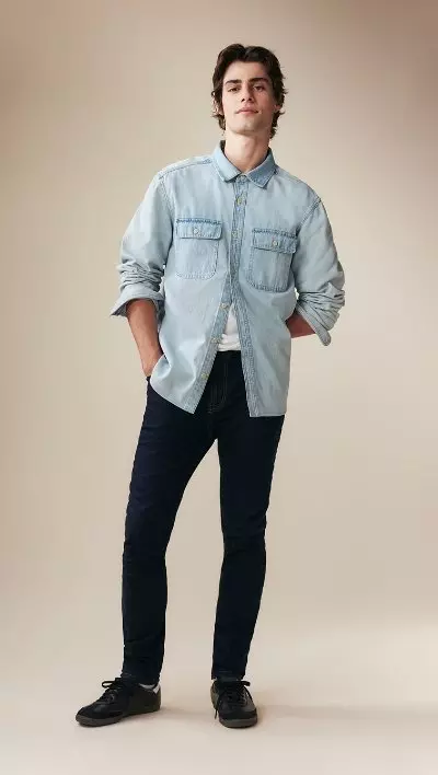 A male model wearing skinny jeans and a light denim button-down shirt.