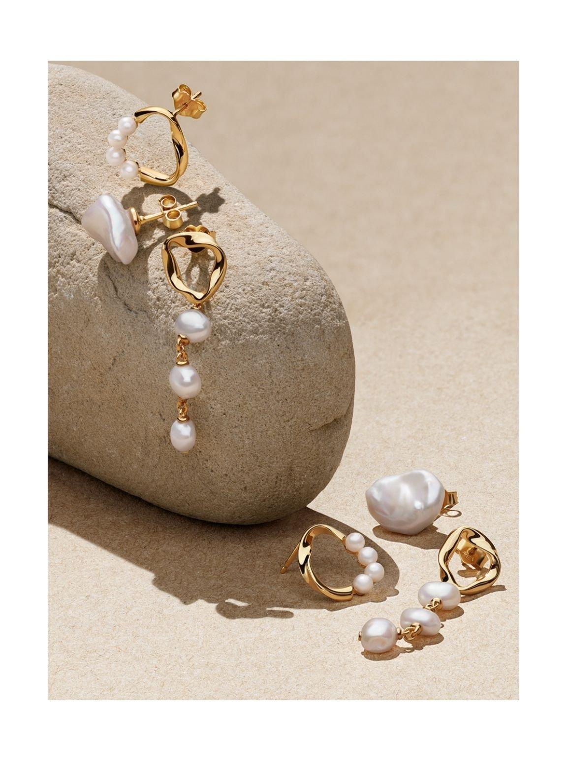 Gold pearl earrings by Pandora lying on a beige stone on sand