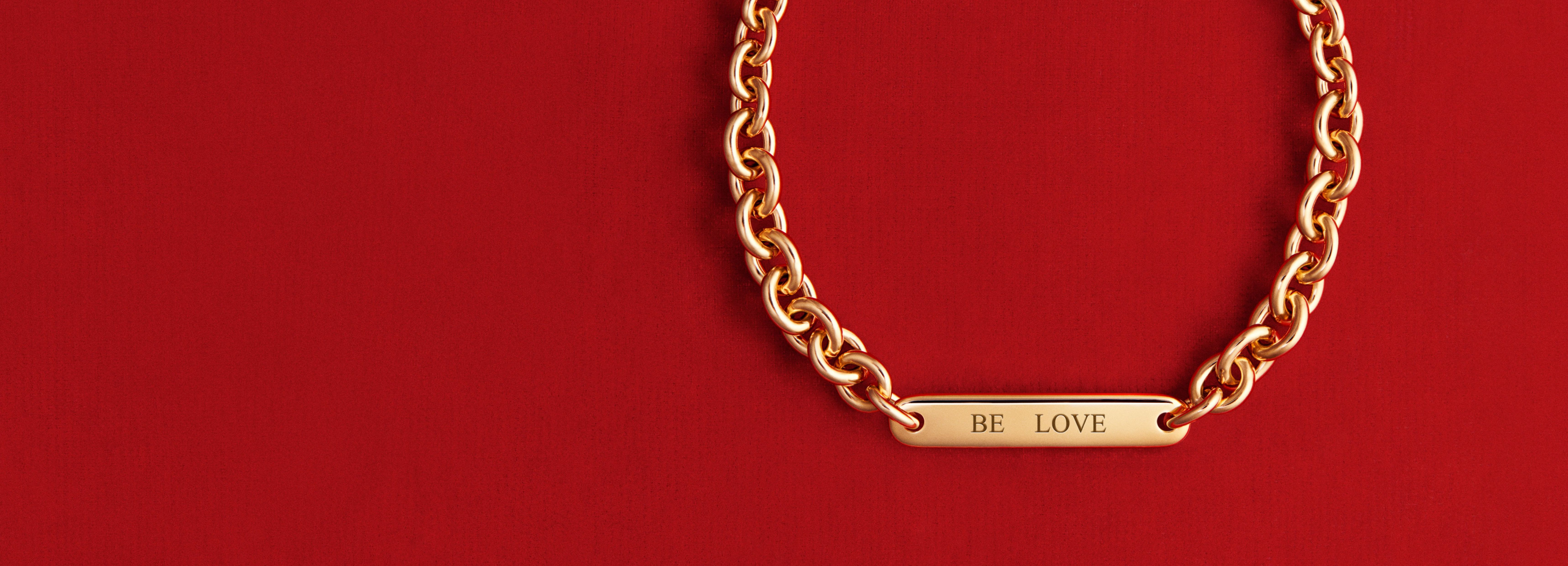 Against a red background hangs a gold Bar Link Bracelet with "Be Love" engraved on the rectangular pendant.