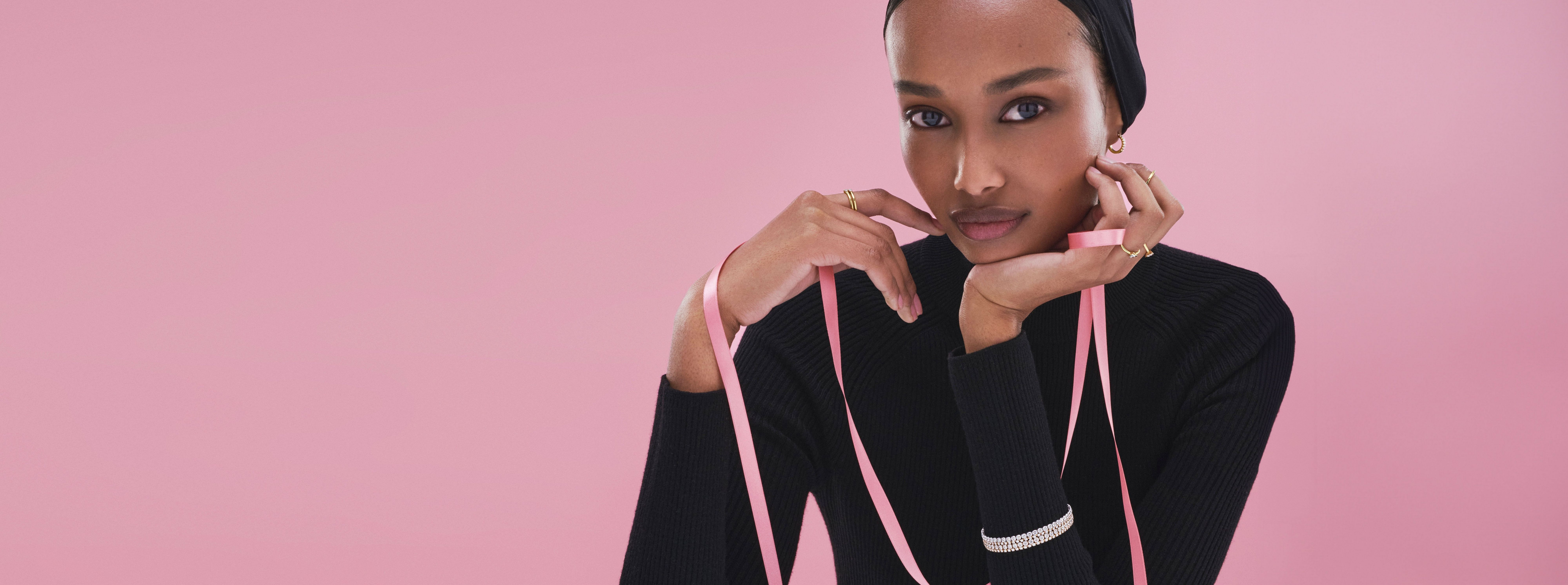 A model rests her head on her left palm, wearing a petite lab-grown diamonds bracelets on her left wrist. She's wrapped in pink Pandora gift ribbons.