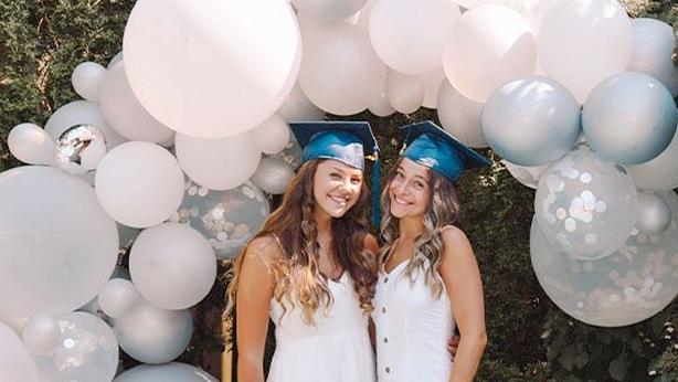 Two high school senior girls wearing white sundresses and blue graduation caps standing outside under a large balloon arch created with various white and silver round latex balloons.