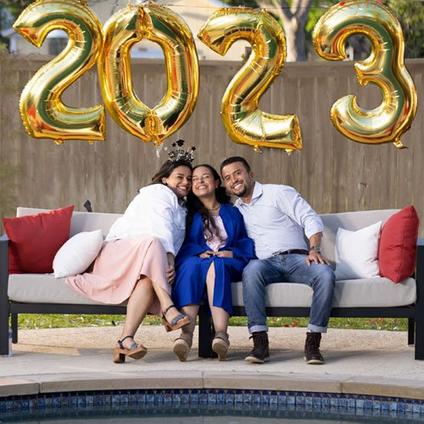 Mom, Graduate Daughter, and Dad Posing for Photo While Sitting on Patio Under Large 2023 Foil Balloons