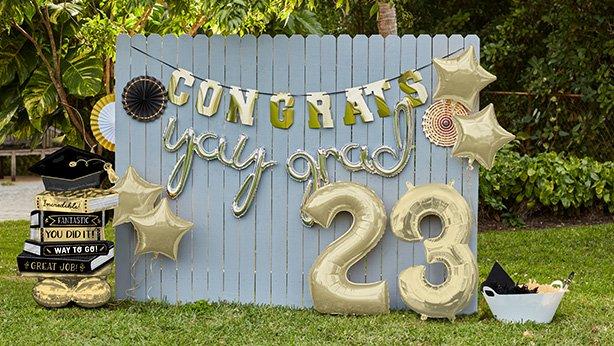 Outdoor photo booth made up of large black and gold foil balloons and an assortment of graduation signs hung on a gray fence wall.