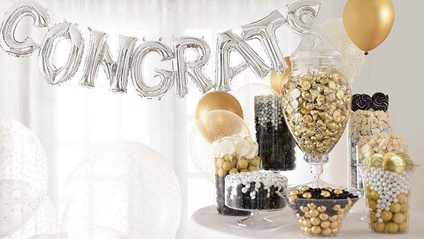 Large glass jars and bowls filled with various black and gold candy surrounded by large black and gold graduation balloon bouquets.