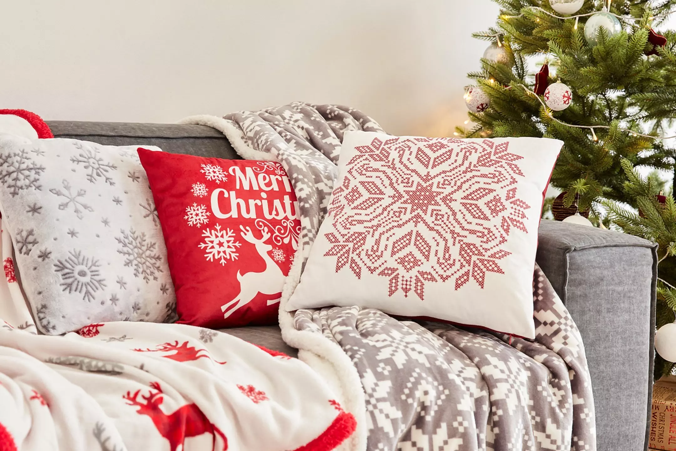 Pepco festive cushions and throws on a gray sofa, featuring red and white Christmas designs next to a decorated tree.