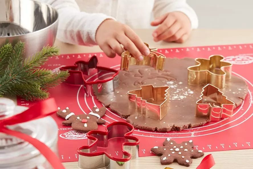 Dziecko wycina świąteczne ciasteczka przy użyciu metalowych foremek od Pepco na czerwonej macie do pieczenia z ciastem piernikowym.