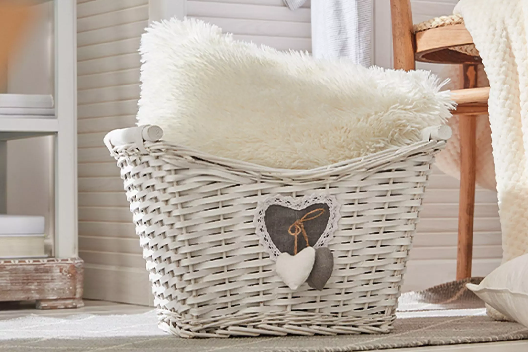 Pepco white wicker basket with heart decoration containing a fluffy cream cushion, displayed in a cozy home setting.