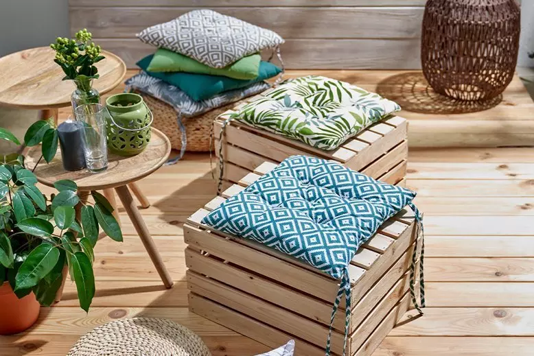 Pepco dekoracyjne poduszki na drewnianych skrzyniach, z wzorami roślinnymi i geometrycznymi, obok stolika z roślinami.