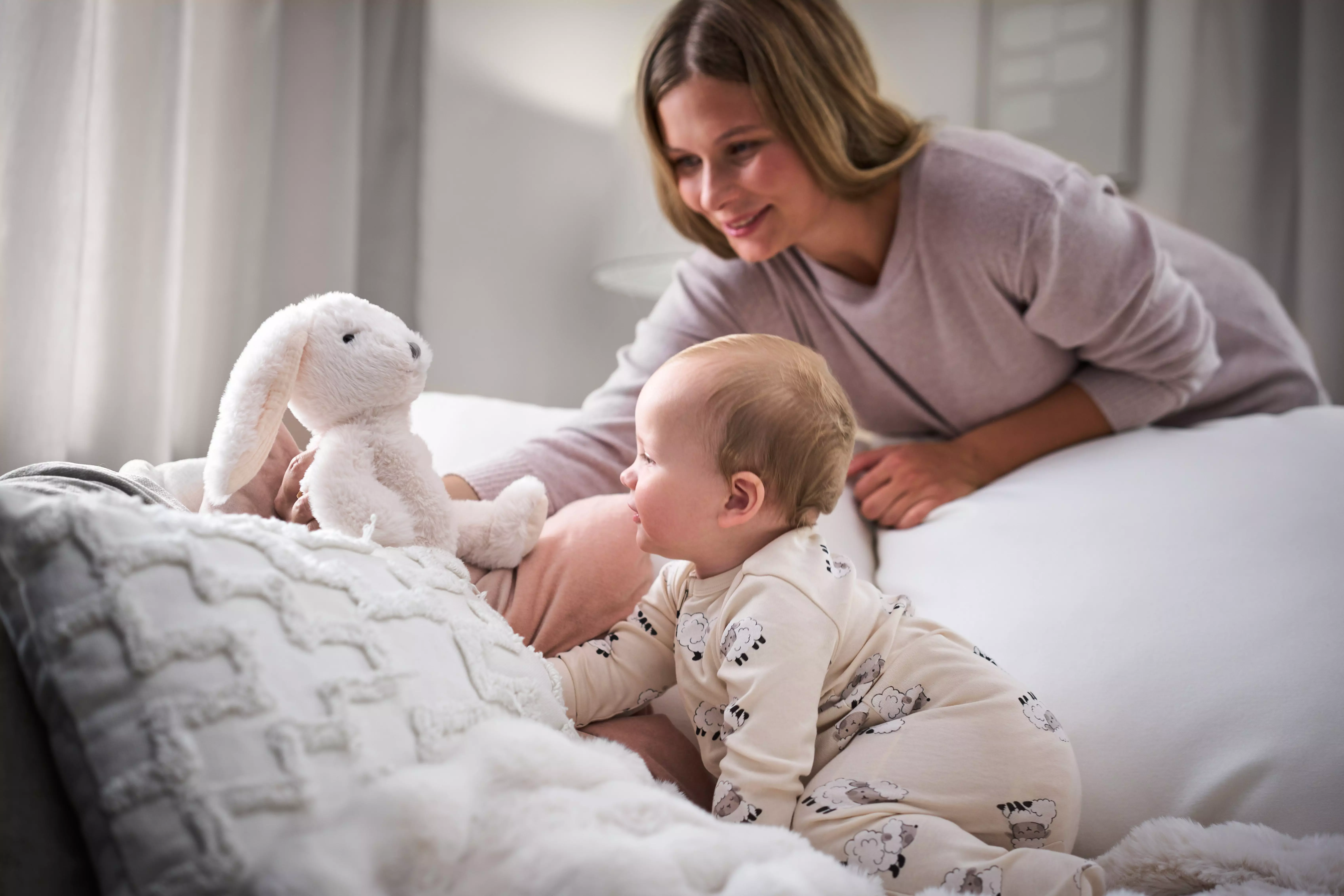 Pepco mother and baby playing with white bunny soft toy on bed in matching beige loungewear.