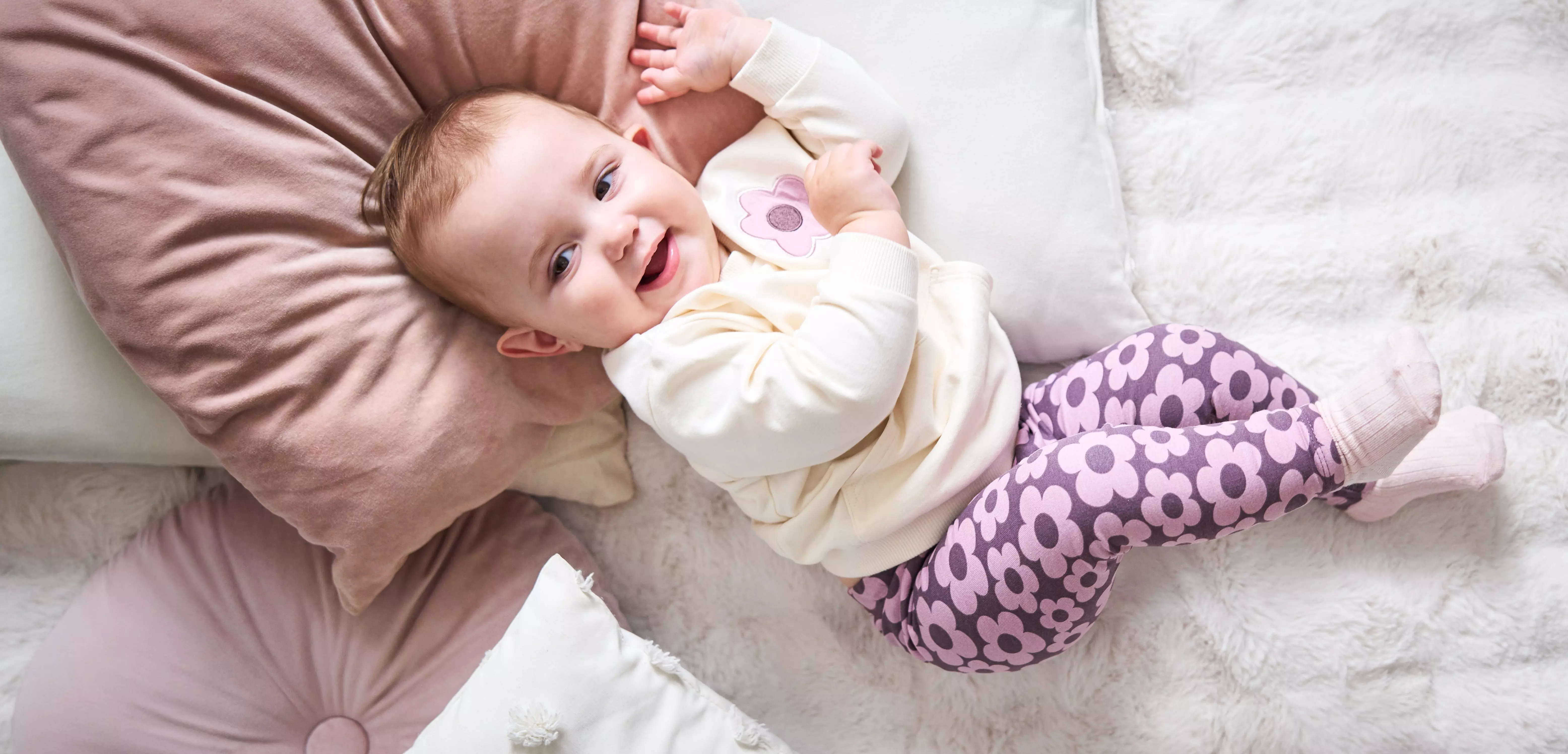 Pepco baby lying on cushion wearing cream top with pink dummy and purple floral leggings.