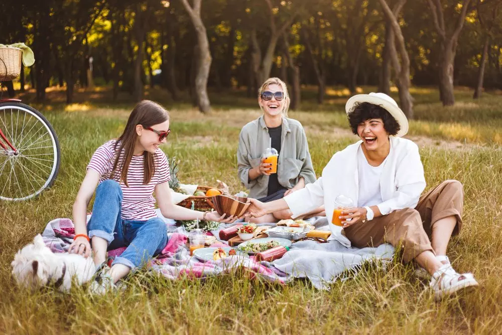 Tre donne fanno un picnic all'aperto su un prato, ridendo e condividendo cibo e bevande.